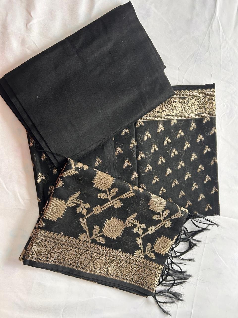 A black Chanderi Suit featuring gold floral and leaf patterns is neatly folded on a white surface. The set includes a matching black blouse piece and a dupatta (pallu) adorned with gold designs and tassels, highlighting the elegance of Chanderi fabric.