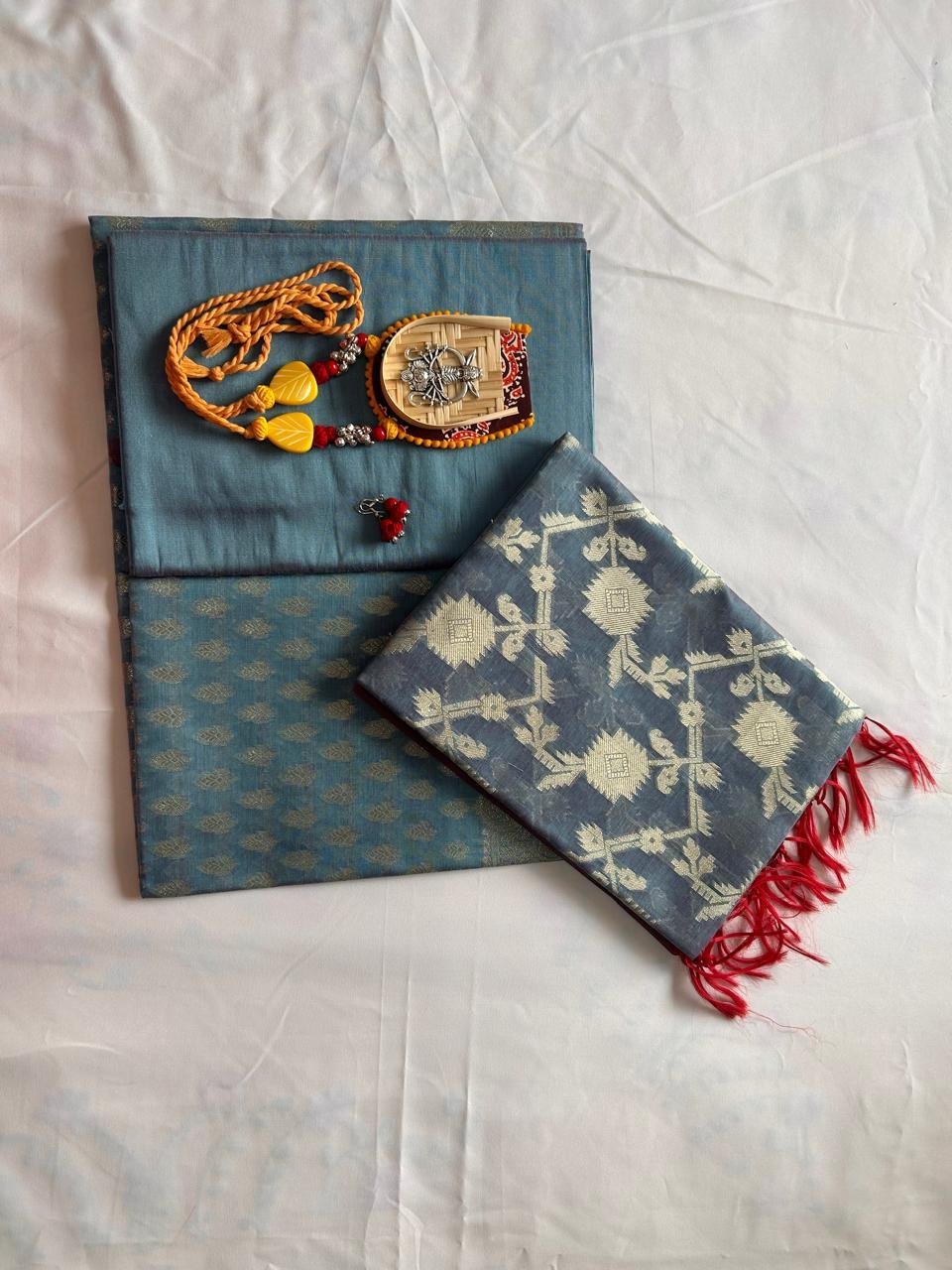 Two folded Chanderi Suits, crafted from luxurious chanderi silk, are displayed—one is adorned with a traditional Indian necklace and a decorative pouch on top. The other suit features elegant white geometric designs and red tassels along the edge, resembling classic saree patterns. Both are beautifully arranged on a light-colored surface.