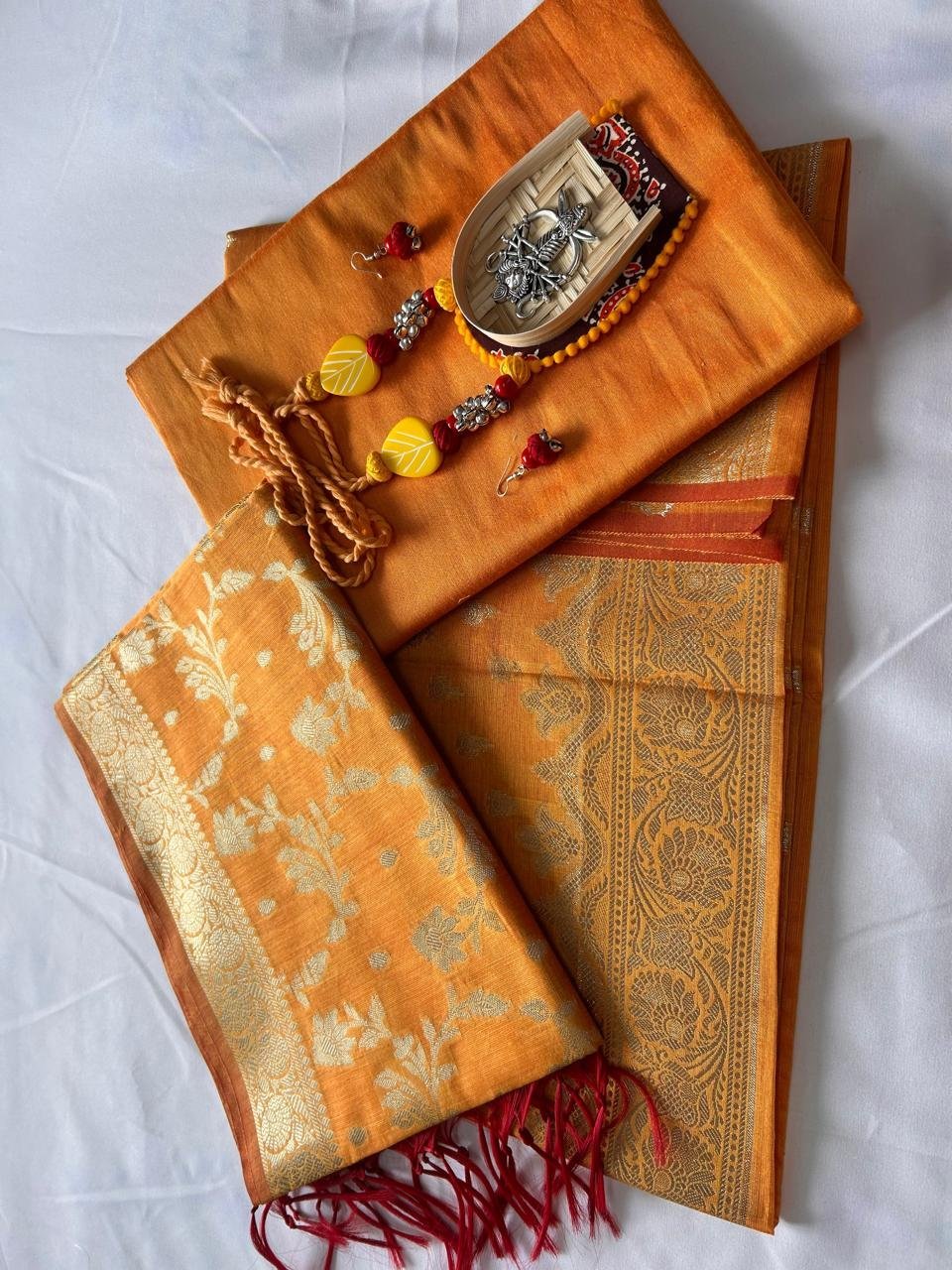 Chanderi Suit in orange Banarasi silk with golden zari work, paired with matching jewelry and wooden boat decor, displayed on a white background.