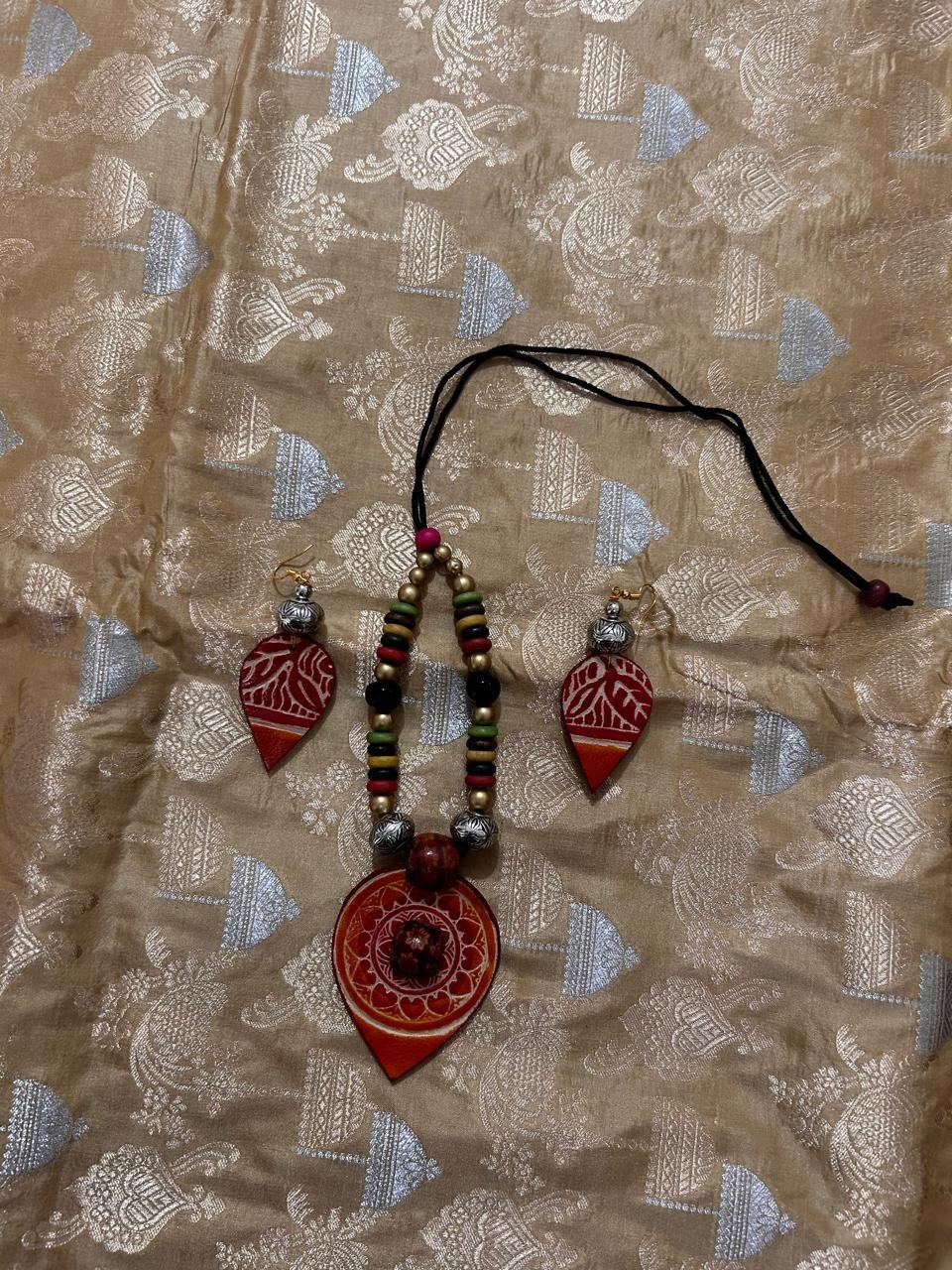 A traditional jewelry set with a pendant necklace and matching earrings in red and brown, displayed on a gold-patterned fabric—perfect for pairing with the Uppada Banarasi Saree.
