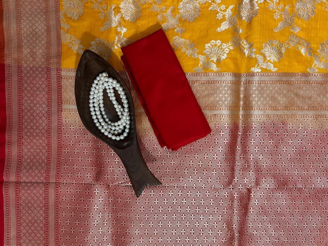 An Uppada Banarasi Saree with red and yellow patterns is spread out, accompanied by a wooden dish holding pearl necklaces and a folded red cloth placed on top.