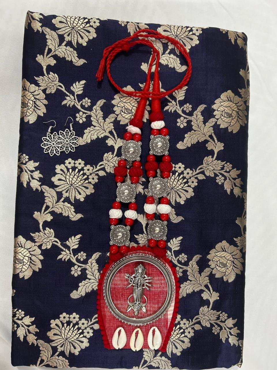 Draped in an Uppada Banarasi Saree in deep blue silk adorned with gold floral motifs, accentuated by a statement red and silver beaded necklace featuring shells and a round pendant detailed with an intricate white design.
